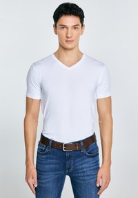 Joven de pie mirando hacia adelante, vistiendo una camiseta blanca lisa de cuello en V, jeans azules y un cinturón marrón, sobre un fondo gris claro.