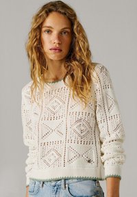 Mladá žena s dlouhými kudrnatými vlasy, oblečená v bílém pleteném svetru s geometrickými vzory a světle modrých džínách na jednobarevném pozadí.