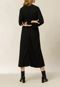 Schwarzes Midikleid mit langen Puffärmeln, taillierter Passform und gerafften Details auf der Rückseite. Kombiniert mit schwarzen Stiefeletten und einer glatten Textur.