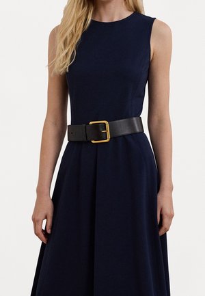 Vrouw draagt een mouwloze marineblauwe jurk met een brede zwarte riem voorzien van een grote gouden rechthoekige gesp.