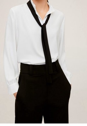 Woman wearing a white long-sleeve blouse with a ruffled collar and a loose black necktie paired with high-waisted black trousers.