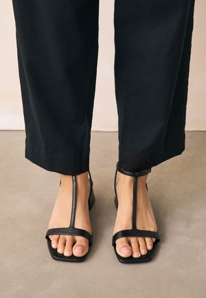Füße tragen schwarze T-Riemchen-Sandalen mit offenen und quadratischen Zehen, kombiniert mit schwarzen kurzen Hosen auf einem neutralen Boden.