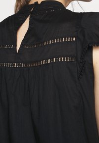 Blusa negra con detalles de encaje y abertura en forma de llave en la parte posterior del cuello, usada por una persona de piel clara y cabello castaño recogido.