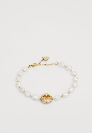 Guldbelagt armbånd med uregelmæssige hvide perleperler, en central gylden cirkulær vedhæng med hul i form af et hjerte, og en lås med et mærke.