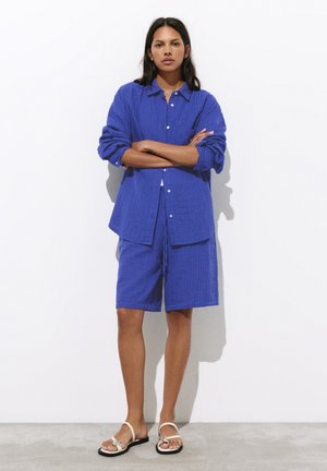 Donna in piedi con le braccia incrociate che indossa una camicia blu strutturata con bottoni e pantaloncini coordinati fino al ginocchio con sandali bianchi.