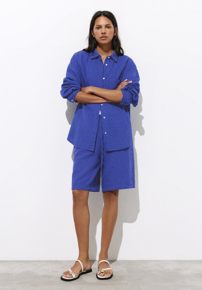 Femme debout, bras croisés, portant une chemise bleue texturée à boutons et un short assorti au niveau des genoux avec des sandales blanches.