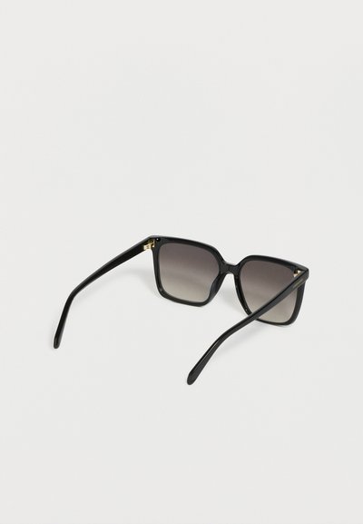Lunettes de soleil noires à monture carrée avec verres dégradés foncés et branches fines, présentées sur une surface blanche.