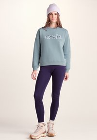 Light blue sweatshirt with "Après" in floral print, paired with dark purple leggings and beige hiking boots. Model wears a light purple beanie.