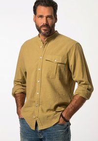 Camicia khaki con collo alla mandarino, maniche lunghe arrotolate fino ai gomiti, tasca frontale e tessuto testurizzato. Indossata con jeans blu.