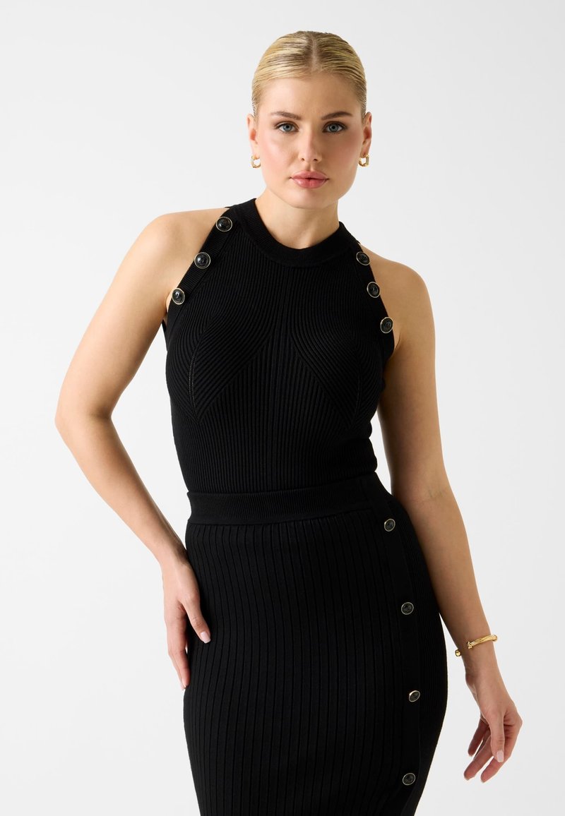 Frau mit blondem Haar, die ein ärmelloses schwarzes, geripptes Kleid mit Knopfdetails an Schultern und Seite trägt, posiert vor weißem Hintergrund.