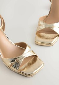 Talons ouverts métalliques dorés avec des brides croisées et un design à bout carré sur une surface gris clair.