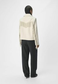 Chaqueta blanca con flecos y cuello, diseñada con acentos de flecos verticales en la parte posterior y combinada con pantalones oscuros de pierna ancha.