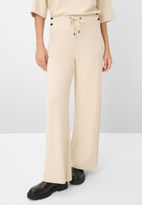 Pantaloni beige a gamba larga realizzati in tessuto liscio, con un cinturino in vita regolabile con dettagli neri. Indossati con stivali platform neri.