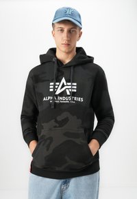 Joven con una sudadera con capucha de camuflaje negro que lleva el logo "Alpha Industries Knoxville, Tennessee" y una gorra de color azul claro, de pie con las manos en los bolsillos de la sudadera.
