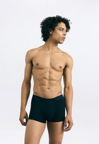 Boxeri scurți din bumbac negru, cu o bandă moale la talie pe care este scris "Calvin Klein"; modelul are o constituție musculară și păr creț, stând pe un fundal deschis.