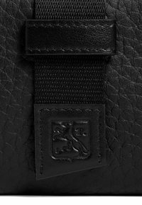 Borsa in pelle nera con superficie texturizzata, una tracolla intrecciata e una targhetta con il logo di un simbolo di leone. I dettagli di cucitura sono visibili.