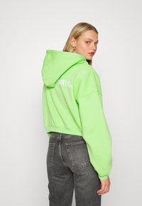 Camisola verde corta com bolsos laterais e capuz com cordão, apresentando texto branco nas costas. Usada com jeans cinza escuro.