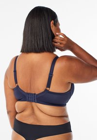 Marineblå bh med tekstureret stof, justerbare stropper og en hægte-lukning på bagsiden. Sort bikiniunderdel synlig.