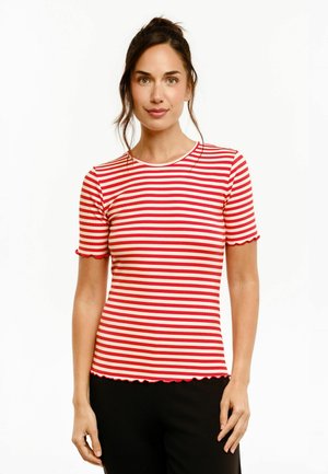 Femme aux cheveux bruns en chignon, portant un t-shirt à manches courtes rayé horizontalement rouge et blanc et un pantalon noir, debout sur fond blanc.