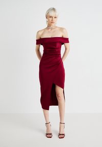 Robe bordeaux dénudée des épaules en tissu lisse, avec des détails froncés et une fente à l'avant. Complète avec des sandales à talons hauts à lanières.
