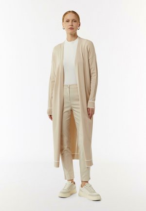 Lange beige cardigan met open voorkant, geribbelde manchetten, gecombineerd met bijpassende beige slim-fit broek en witte sneakers. Gladde textuur, minimalistisch ontwerp.