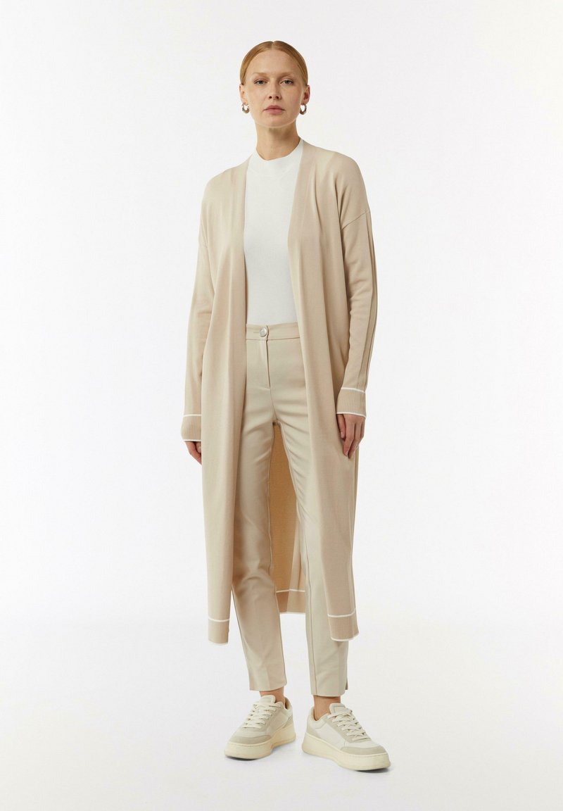 Langer beiger Cardigan mit offener Vorderseite, gerippten Bündchen, kombiniert mit passenden beigen, eng anliegenden Hosen und weißen Sneakers. Glatte Textur, minimalistisches Design.