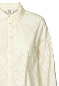 Lichte crème blouse met een textuur van bloemmotieven, klassieke kraag en knoopsluiting aan de voorkant. Lange mouwen met een geplooid detail bij de schouders.
