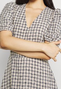 Ärmlös klänning med puffärmar i ljusbeige tyg med marinblå rutmönster. Har en V-ringning och en rynkad midja för extra textur.