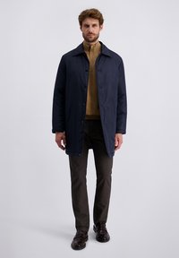 Manteau long bleu marine avec fermeture à boutons, poches latérales et texture lisse, associé à un pull à col haut beige et un pantalon marron à motifs.