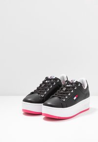 Černé kožené tenisky s bílou platformou a jasně růžovou podešví. Mají šněrování v přední části, logo a hladkou texturu.