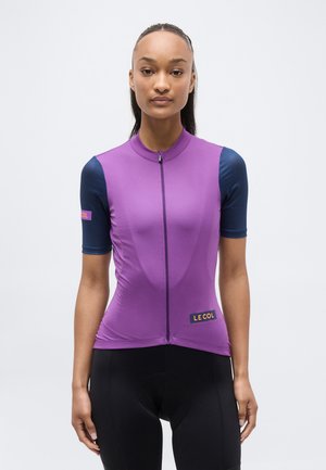 Vrouw draagt een paarse wielertrui met donkerblauwe mouwen, ritssluiting aan de voorkant en "LE COL" logo's op borst en mouw.