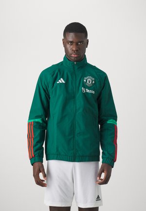 Chaqueta deportiva verde con cuello alto, con detalles en franjas rojas y verdes en las mangas, logo del Manchester United y cierre con cremallera.