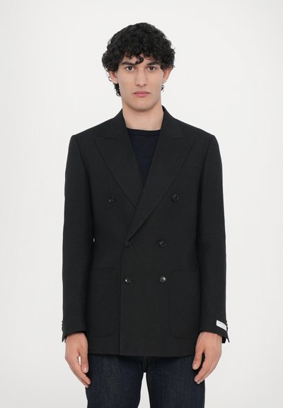 Schwarze, zweireihige Blazer mit taillierter Passform, Reverskragen, zwei vorderen Taschen und subtiler Textur. Getragen über einem dunklen Hemd.