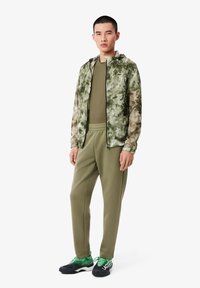Jachetă verde cu camuflaj și fermoar negru; cămașă solidă de măsline și pantaloni jogger asortați; adidași negri cu accente verzi. Material neted, ajustat pe corp.