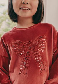 Red plush sweatshirt with a large bow design made of sequins, featuring a soft texture and round neckline.