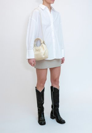 Personne portant une chemise blanche oversize, une mini-jupe beige, des bottes noires montantes et portant un petit sac à main beige avec des doubles poignées.