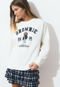 Un sweat-shirt blanc avec le texte "BROWNIE" et un graphique de chien brodé. Associé à une jupe en tartan bleu marine à plusieurs couches avec des détails en dentelle.