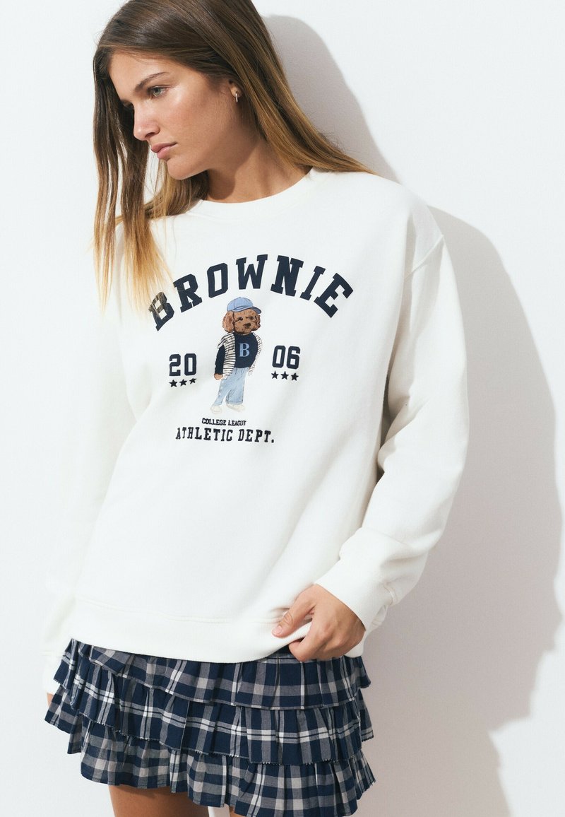 Un sweat-shirt blanc avec le texte "BROWNIE" et un graphique de chien brodé. Associé à une jupe en tartan bleu marine à plusieurs couches avec des détails en dentelle.