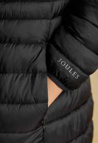 Čierna prešívaná bunda s mäkkou textúrou a viditeľným zipsom. Na manžete je vytlačené logo "JOULES".