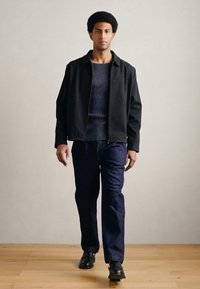 Čierna bunda so zipsom cez tmavomodrý rebrovaný sveter, spolu s tmavými nohavicami a čiernymi topánkami. Model stojí na drevenej podlahe pred jednoduchou stenou.