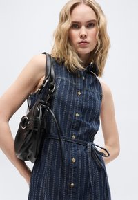 Ξανθιά γυναίκα που φοράει αμάνικο πουκαμισοφόρεμα από τζιν με κουμπιά και ζώνη, κρατώντας μαύρη δερμάτινη τσάντα ώμου σε λευκό φόντο.