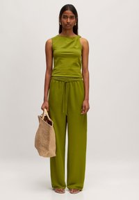 Groene tweedelige outfit met een mouwloze top en ontspannen, met een koord verstelbare broek, gemaakt van zachte, gestructureerde stof. Model houdt een geweven tas vast.