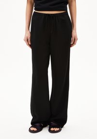 Svarta vida byxor med elastisk midja och snöre. Slät tygtextur, hel längd, parat med mörka platta sandaler.