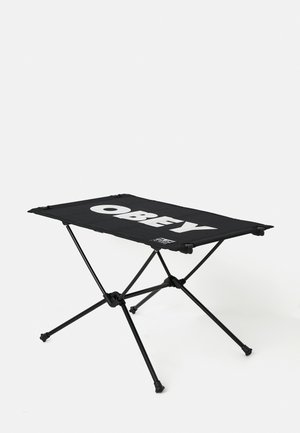 Schwarzer tragbarer Campingtisch mit einer flachen Oberfläche, auf der das Wort "OBEY" in fetter weißer Schrift dargestellt ist, gestützt von einem klappbaren Metallrahmen.