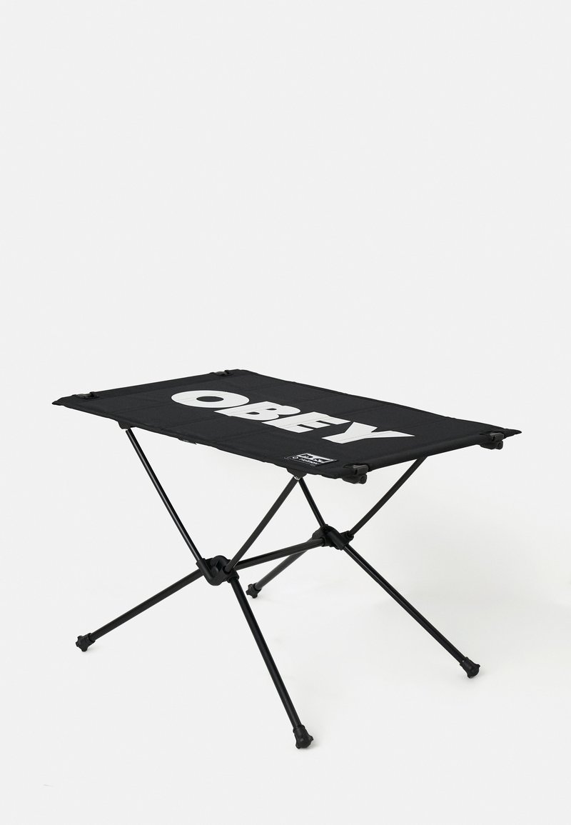 Zwarte draagbare kampeertafel met een vlakke oppervlakte waarop het woord "OBEY" in vetgedrukte witte letters staat, ondersteund door een opvouwbaar metalen frame.