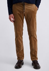 Man wearing brown corduroy pants with drawstring waist and dark brown leather loafers, standing with hands in pockets.