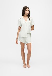 Jeune femme debout pieds nus portant une chemise légère à manches courtes boutonnée et un short assorti sur un fond blanc uni.