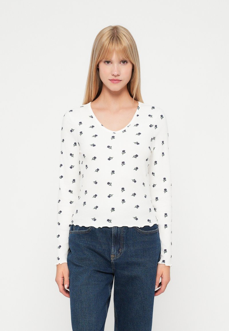 Λευκή μπλούζα με μακριά μανίκια και ριπ ύφανση, διαθέτει σκαλπμένο στρίφωμα, V-λαιμόκοψη και μικρά σκούρα μπλε λουλούδια σε όλη την επιφάνεια.