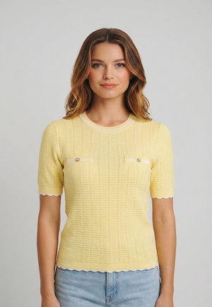 Mujer con cabello castaño claro que lleva un suéter amarillo de manga corta con botones decorativos y jeans azul claro, frente a un fondo gris liso.