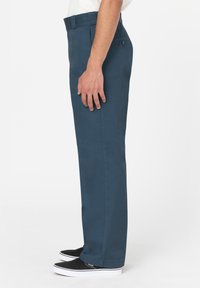 Boční pohled na osobu v tmavě modrých rovně střižených kalhotách a černých slip-on teniskách na bílém pozadí.